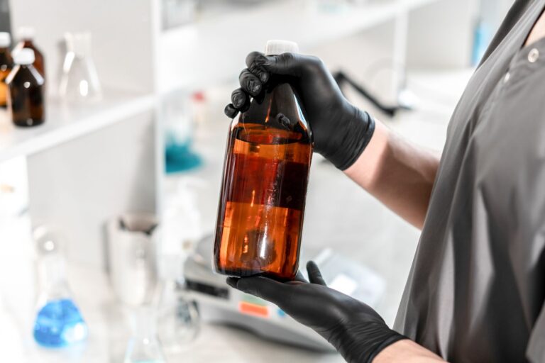 Manos con guantes negros sosteniendo un frasco de vidrio ámbar en un laboratorio. Foto: Ivan S, Pexels